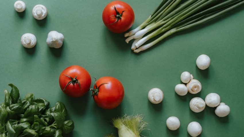 Flat lay of fresh vegetables including tomatoes, mushrooms, and spring onions on a green surface.